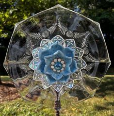 a blue flower is in the middle of a clear glass stand on some grass with trees in the background