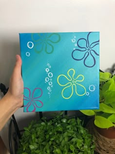 a hand holding up a painting with flowers and bubbles on it, in front of some potted plants