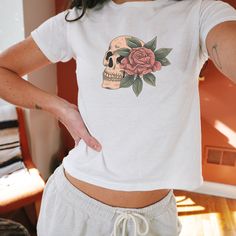 Introducing our Women's Vintage Skull & Rose Baby Tee, a unique blend of edgy and elegant. This stylish baby tee features a striking design of a skull adorned with a massive, intricately detailed rose covering one side of the face. The vintage-inspired print adds a touch of retro charm, making it perfect for those who love to mix classic aesthetics with a modern twist. Crafted from soft, high-quality fabric, this tee ensures a comfortable fit while making a bold fashion statement. Whether you're dressing it up or down, this tee is a versatile addition to any wardrobe, ideal for expressing your individual style with a hint of vintage allure. White Cotton T-shirt With Rose Print, Cotton Graphic Tee With Rose Print, Cotton Crew Neck Top With Rose Print, White Cotton Tops With Rose Print, Fitted Cotton Top With Rose Print, White Cotton Top With Rose Print, White Cotton Rose Print Top, Fitted Graphic Tee With Skull Print, Pink Halloween Graphic Tee