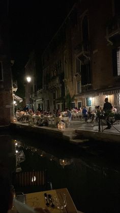 people sitting at tables on the side of a river in front of buildings and lights