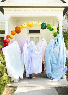 three people dressed in ghost costumes standing on the front porch of a white house with pumpkins hanging from it