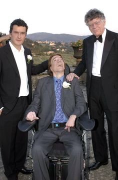 three men in tuxedos standing next to each other with one man in a wheel chair