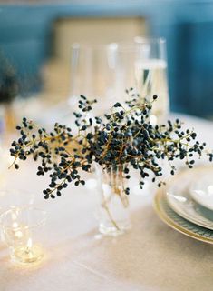 an arrangement of black berries in a vase on a white table cloth with place settings