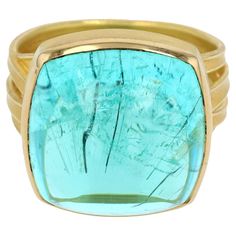 Experience the extraordinary with this captivating 18K yellow gold ring by Barbara Heinrich, featuring a stunning 26.20-carat cushion-shaped Mozambique Paraiba tourmaline cabochon. The large, vibrant tourmaline, with its enchanting teal hue, is the centerpiece of this exquisite piece, exuding a luminescent glow that captivates the eye. Set on a unique 4-ribbon band, the design harmonizes contemporary elegance with timeless sophistication. The band’s fluid lines and polished finish enhance the gem’s brilliance, making it a truly mesmerizing statement piece. This ring is not only a testament to the beauty of the rare Paraiba tourmaline but also to the exceptional craftsmanship and artistry of Barbara Heinrich. Barbara Heinrich, a renowned jewelry designer, is celebrated for her meticulous at