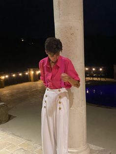 a woman standing next to a pillar in front of a swimming pool wearing pink shirt and white pants