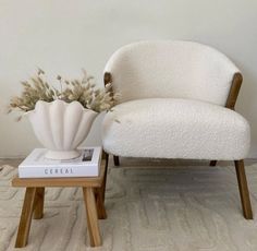 a white chair sitting next to a table with a book on it and a vase filled with flowers