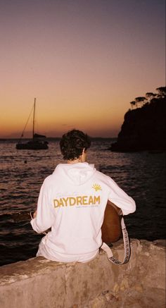 a man sitting on the edge of a stone wall next to the ocean with a sailboat in the background