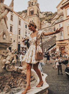 a woman standing in front of a fountain with her hand out to the side and people walking around