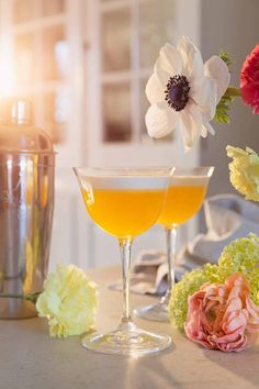 two glasses filled with drinks sitting on top of a table next to flowers and a cocktail shaker