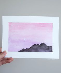 a hand holding up a watercolor painting with mountains and the moon in the sky