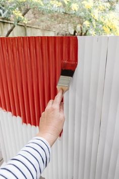 a person with a paintbrush painting a red and white fence outside on a sunny day