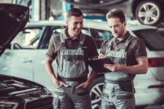 two men in overalls looking at something on a clipboard next to a car