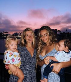 three women and one baby are posing for a photo in front of the water at sunset