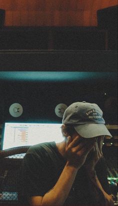 a man sitting in front of a computer screen talking on a cell phone while wearing a hat