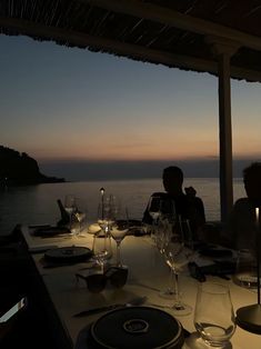 two people sitting at a table with wine glasses and plates on it in front of the ocean