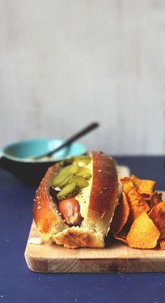 a hot dog with pickles and chips on a cutting board next to a drink