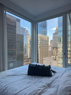 a black bag sitting on top of a white bed in front of two large windows