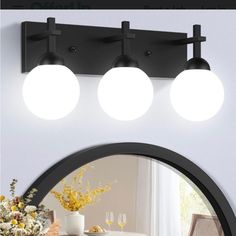 three lights on the wall above a mirror in front of a table with flowers and wine glasses