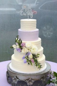 a white wedding cake with purple flowers and greenery on the top is sitting on a table