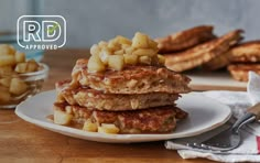 a stack of pancakes topped with apples on top of a white plate next to bananas