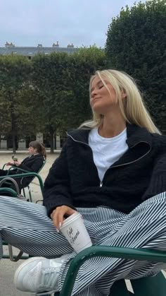 a blonde woman sitting on top of a green chair next to a park bench with trees in the background