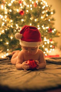 a baby wearing a santa hat playing with a red ornament in front of a christmas tree