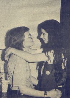 two women sitting next to each other in front of a white wall and one is hugging the other