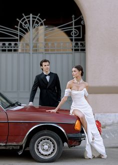 a young man and woman standing next to a red car in front of a building