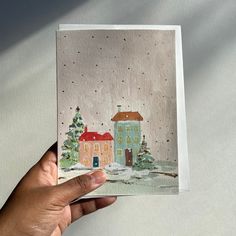 a hand holding up a card with a house and trees on it in the snow