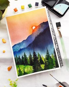 watercolor landscape painting on paper with paintbrushes next to it and other art supplies