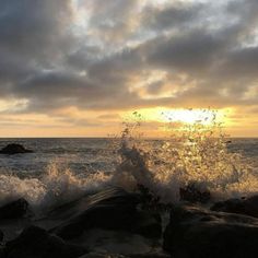 the sun is setting over the ocean with waves crashing on rocks