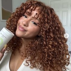 a woman with curly red hair holding a tube of cream on her face and looking at the camera