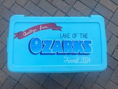 a blue lunch box sitting on top of a brick floor next to a sidewalk with the words lake of the ozarks printed on it