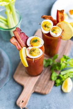 two cocktails with bacon, hard boiled eggs and garnishes on a cutting board