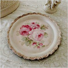 a plate with pink roses painted on it sitting on a lace tablecloth next to a vase