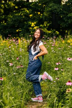 a woman in overalls and pink shoes standing in a field full of wildflowers