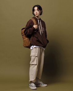 a young man standing in front of a brown wall wearing a brown sweatshirt and khaki pants