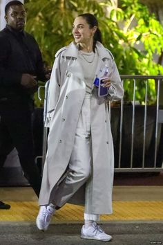 a woman in grey trench coat and white sneakers walking down the street with a water bottle