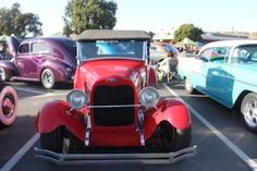 an old red car parked in a parking lot next to other antique cars and trucks