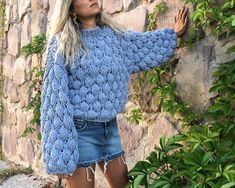 a woman leaning against a stone wall with her hands in the air and wearing a blue sweater