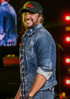 a smiling man wearing a denim jacket and hat standing in front of a large screen