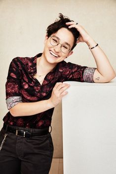 a man with glasses leaning against a white box smiling at the camera while wearing black pants and a red shirt