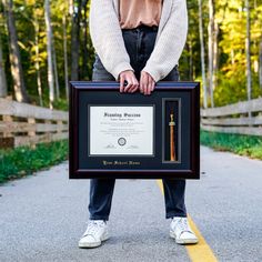 a woman holding up a framed award on the side of a road with trees in the background