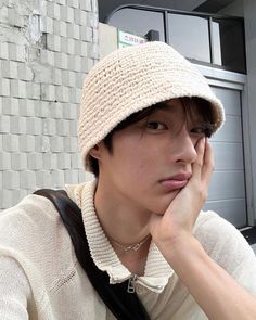 a young man wearing a white hat and holding his hand to his face while sitting in front of a brick wall