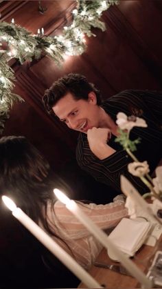 a man and woman sitting at a table with candles in front of them, looking at each other