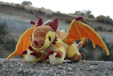 a stuffed toy dragon sitting on top of a rock covered ground with trees in the background