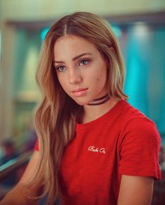 a young woman with long hair wearing a red t - shirt and looking at the camera