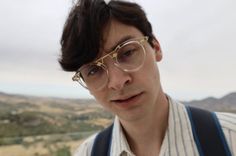 a young man wearing glasses and suspenders looking at the camera with mountains in the background