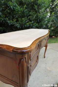 an old wooden desk sitting on top of a cement floor next to some bushes and trees