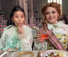 two women sitting at a table with food and drinks in front of them, posing for the camera